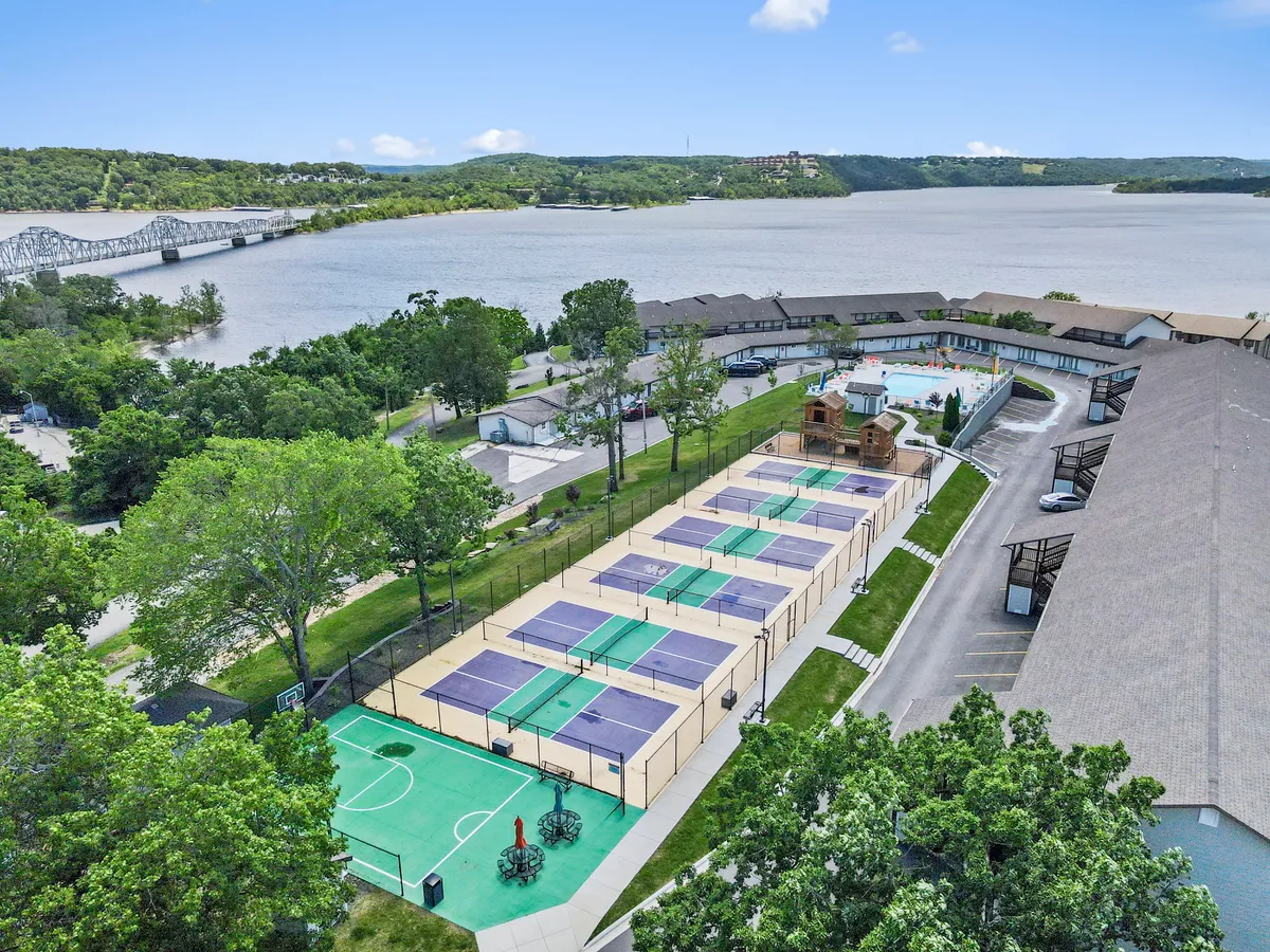 A vibrant outdoor scene showcasing tennis courts, a basketball area, a swimming pool, lush surrounding greenery, and serene waterfront views. The courts are set up for play and the pool is ready for family fun, with ample space for various activities.