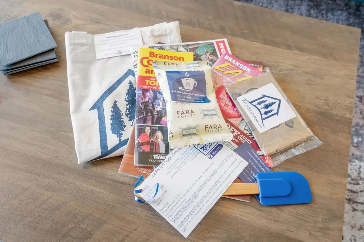 An inviting arrangement featuring a branded tote bag, brochures, coffee packages, and a blue spatula on a wooden surface. The image suggests guests can plan their outings, enjoy local flavors, and receive helpful information about Branson attractions.