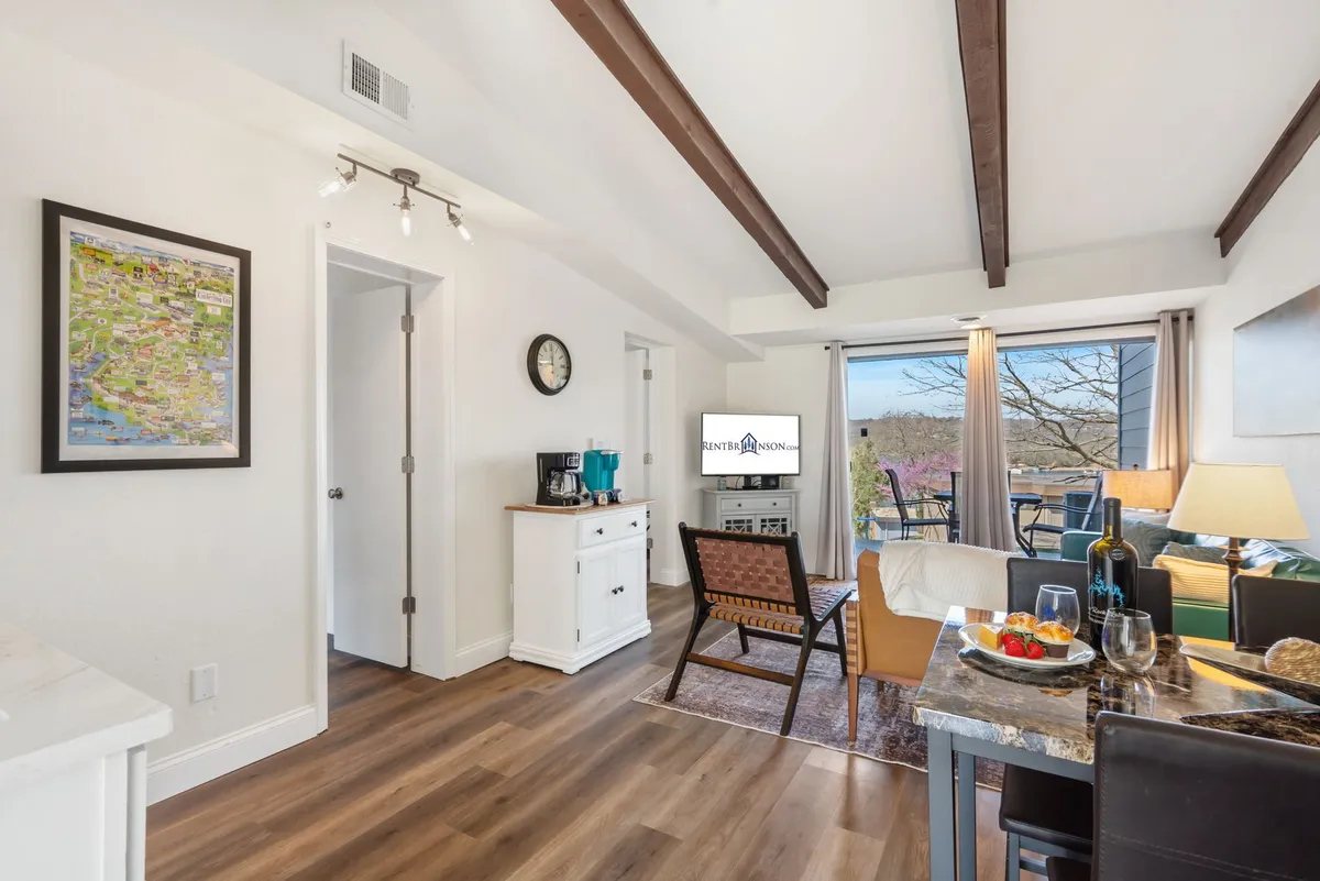 A warmly lit open-concept living and dining area, featuring a comfortable couch with a throw blanket, a dining table topped with a bowl of fruit, modern wall art, and a prominent Smart TV. The setting invites both relaxation and social gatherings, with enough space for families or couples.