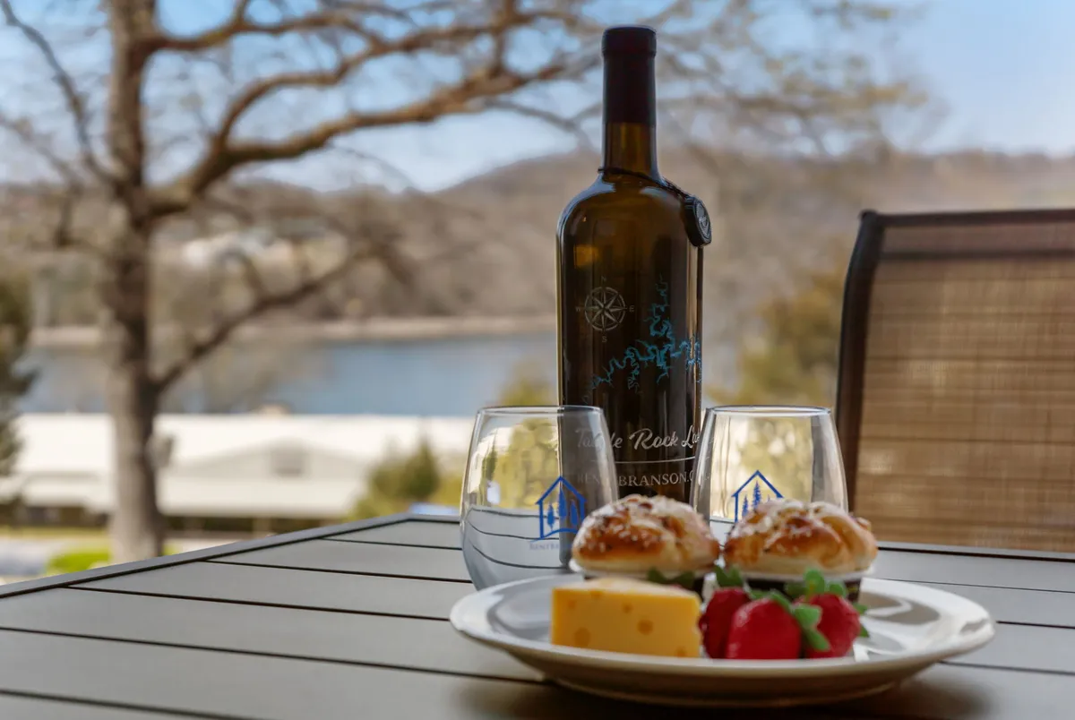 A serene outdoor setting features a wine bottle and glasses on a table, accompanied by a food platter. The scene overlooks a tranquil lake, with gentle hills and greenery in the background, creating an inviting atmosphere for relaxation and shared experiences.