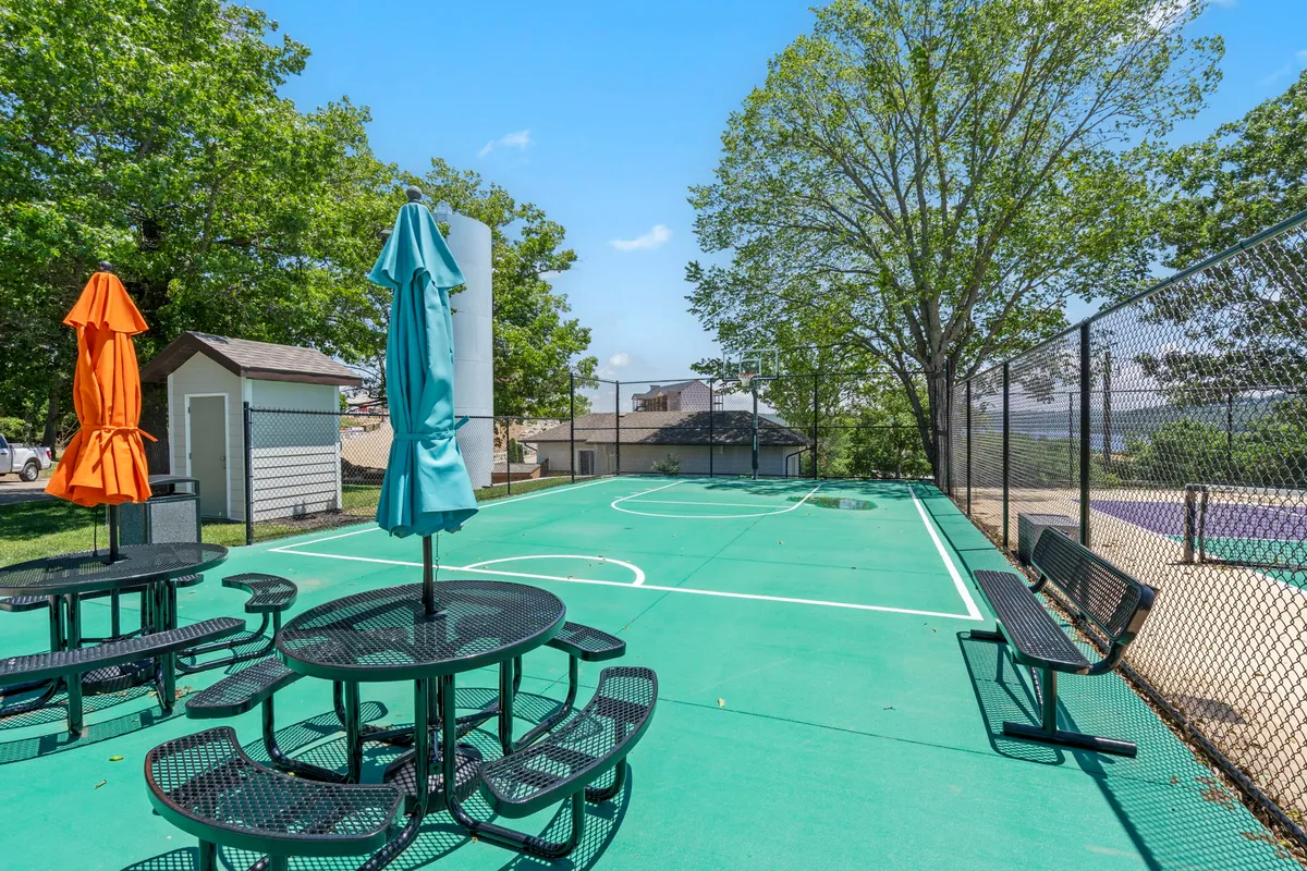 An energetic exterior scene featuring a basketball court surrounded by lush greenery, colorful umbrellas, and picnic tables. The space is set up for basketball games, casual social gatherings, and outdoor relaxation, all under the shade of mature trees.